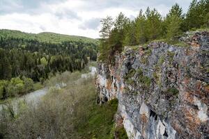 rochoso montanhas do cinzento cor esticam ao longo a rio, uma Alto penhasco, uma montanha paisagem, uma conífero floresta cresce em a montanha, atrasado verão. foto