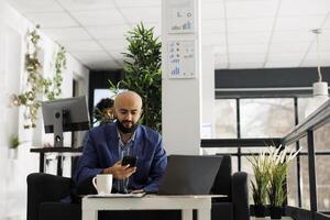 o negócio árabe profissional mensagens de texto em Smartphone enquanto trabalhando em computador portátil dentro começar acima escritório. empreendedor procurando financeiro em formação em Móvel telefone dentro verde aberto espaço foto