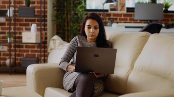 auto empregado mulher focado em acabamento maçante monótono trabalho tarefas enquanto ficando às casa dentro agradável apartamento. freelancer empresária digitando em computador portátil teclado, descansado em sofá, Câmera b panning tiro foto