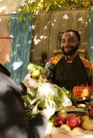 alegre agricultor apresentando localmente crescido maçãs em agricultores mercado ficar em pé, pequeno o negócio orgânico produtos. africano americano homem falando para Senior cliente, vários fresco frutas e legumes. foto