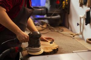 carpinteiro segurando poder ferramenta usava para suave superfícies de abrasão com lixa, usando isto em madeira pedaço. homem usando orbital lixadeira equipamento para mobília montagem trabalho dentro estúdio foto