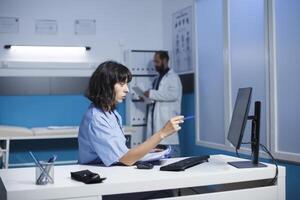 determinado caucasiano enfermeira verificação e analisando dela notas às a clínica escritório escrivaninha. jovem mulher vestindo azul esfrega preparando para médico consultas com pacientes. foto