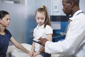 Preto homem vestindo laboratório casaco consulta com menina paciente e mãe dentro clínica escritório. africano americano médico usando digital tábua enquanto fornecendo médico adendo para caucasiano mulher e dela filha. foto