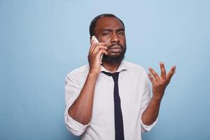 franzindo a testa masculino trabalhador autonomo explicando problema em Smartphone fazendo mão gesto contra azul fundo. chateado africano americano homem de negocios dentro mau humor tentando para Compreendo o negócio conversação. foto