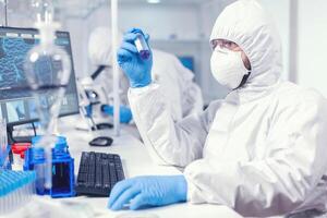 laboratório técnico vestido dentro protetora terno Como segurança precaução olhando às teste tubo. cientista dentro laboratório vestindo macacão fazendo pesquisa e analisando substância durante global pandemia com COVID-19. foto