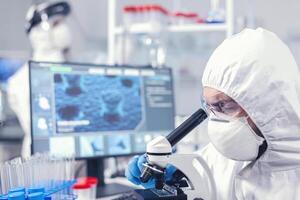 médico equipe dentro laboratório conduzindo coronavírus análise usando microscópio. cientista dentro protetora terno sentado às local de trabalho usando moderno médico tecnologia durante global epidemia. foto