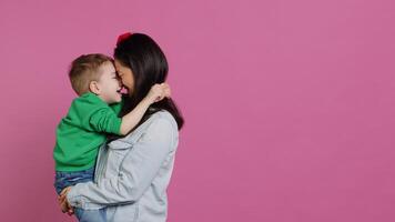 adorável pequeno Garoto abraçando dele mãe e jogando em volta, tendo Diversão contra Rosa fundo. jovem criança pequena posando com dele mãe segurando ele, rindo e ser alegre. Câmera uma. foto