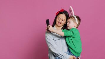 adorável pequeno criança levando As fotos com dele mãe em Smartphone, tentando para capturar Diversão e fofa momentos contra Rosa fundo. pequeno Garoto ser brincalhão e enganar por aí com telefone. Câmera uma.