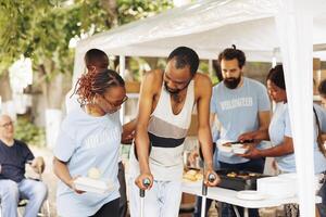 multiétnico caridade grupo entrega Fora livre Comida e nutrição para Menos afortunado e desabilitado. retrato do Preto mulher com azul camiseta ajudando a pobre, carente africano americano homem em muletas. foto