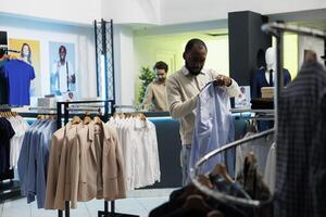 africano americano comprador verificação Tamanho em camisa rótulo enquanto escolhendo na moda formal vestem dentro compras Shopping boutique. jovem homem navegando elegante vestuário em cabide dentro sala de exposições foto