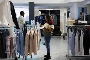 mulher segurando camisa em cabide e examinando estilo enquanto compras dentro boutique. jovem africano americano comprador procurando formal vestem e escolhendo na moda vestuário dentro roupas loja foto