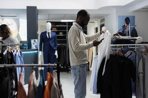 varejo mercado boutique cliente examinando suspensão vestuário em prateleira enquanto selecionando Tamanho e estilo dentro Smartphone aplicativo. homem escolhendo vestuário dentro roupas loja e verificação desconto em Móvel local na rede Internet foto