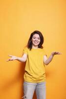 alegre jovem mulher com expressivo positivo atitude acolhedor com elevado braços dentro estúdio. retrato do morena senhora sorridente, ser amigáveis e sociável, fazendo uma cumprimento gesto. foto