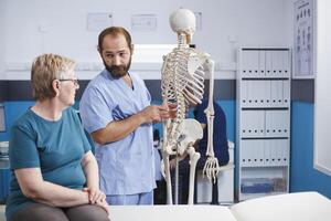enfermeira demonstra espinhal cordão em humano esqueleto para idosos mulher dentro fisioterapia clínica. masculino médico discute anatomia e espinhal ossos com aposentado velho paciente sentado em hospital cama. foto