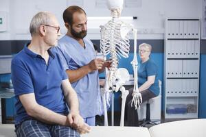 dentro fisioterapia clínica, ortopedista explica espinhal cordão em humano esqueleto para idosos paciente. médico discutindo anatomia e espinhal ossos com velho homem dentro preparação para osteopatia diagnóstico. foto