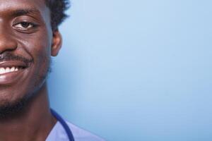 fechar-se retrato do alegre masculino enfermeira vestindo esfrega e estetoscópio em uma azul fundo. metade face, cortada imagem do confiante africano americano médico médico sorridente às Câmera. foto