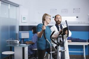 médico consulta com velho paciente fazendo ginástica para recuperação às fisioterapia clínica. envelhecido mulher usa estacionário bicicleta para fisica exercício enquanto falando para uma cuidados de saúde especialista. foto