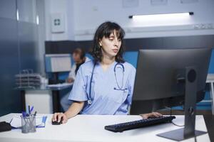 detalhado imagem do uma caucasiano enfermeira operativo uma Área de Trabalho computador dentro uma hospital escritório. uma fêmea praticante vestindo azul esfrega Verificações médico arquivos em uma computador dentro uma clínica sala. foto