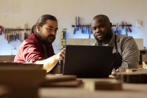 marceneiro olhando sobre plantas em computador portátil, debate com colega Próximo passos dentro madeira em processamento. fabricante e bipoc Aprendiz lendo mobília montagem esquemas dentro estúdio foto