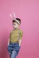 adorável pequeno Garoto vestindo fofo Coelho orelhas e posando com confiança em Câmera, sentindo-me animado sobre Páscoa celebração e em pé contra Rosa pano de fundo. alegre pré-escolar com adorável roupa. foto