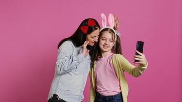 brincalhão alegre mãe e menina sorridente para fotos em Smartphone, tendo Diversão abraçando cada de outros dentro estúdio. pequeno criança a dela mãe posando para fotos, enganar em volta. Câmera b.