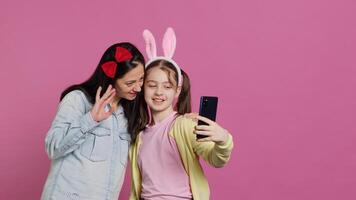 brincalhão alegre mãe e menina sorridente para fotos em Smartphone, tendo Diversão abraçando cada de outros dentro estúdio. pequeno criança a dela mãe posando para fotos, enganar em volta. Câmera uma.