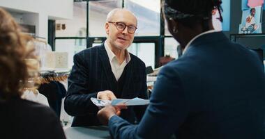 varejo assistentes vendendo roupas para idosos cliente às moda butique, dando desconto cupom para regular cliente. empregados equipe ajudando Senior homem para Comprar roupas Itens em oferta. Câmera b. foto