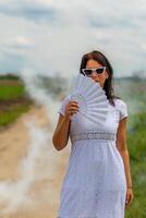 mulher é segurando uma branco ventilador dentro dela mão enquanto caminhando em uma sujeira estrada foto