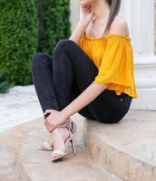modelo vestindo Preto jeans, Alto calcanhares sandálias e amarelo blusa é sentado em concreto escadas foto