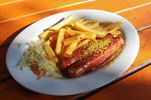 currywurst tradicional alemão, servido com batatas fritas em um prato branco. mesa de madeira como pano de fundo. foto