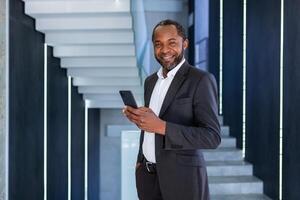 retrato do feliz bem sucedido africano americano chefe, homem sorridente e olhando às Câmera, homem de negocios segurando telefone usando conectados aplicativo, homem caminhando baixa corredor do construção dentro escritório. foto