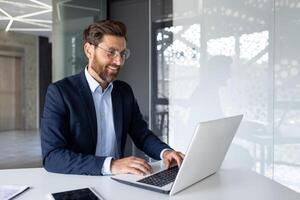 sorridente homem de negocios trabalhando em computador portátil dentro moderno escritório, refletindo produtividade, sucesso e profissional contentamento. foto