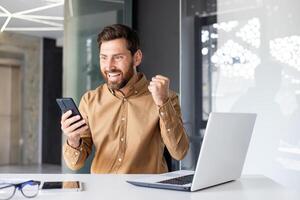 homem vencedora segurando telefone dentro mãos, homem de negocios às local de trabalho feliz com conectados realização resultados, trabalhador usando aplicativo em Smartphone, sentado dentro escritório com computador portátil, vestindo casual camisa. foto