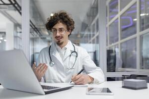 uma médico vestindo uma branco laboratório casaco e estetoscópio sentado dentro frente do uma computador portátil computador. ligar homem sorrir falando foto