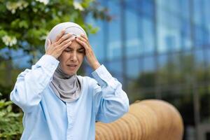 uma profissional mulher dentro uma hijab parece estressado e detém dela cabeça, indicando uma dor de cabeça ou muito pesado sentimentos, com a escritório fundo. foto