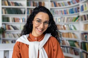 retrato do jovem lindo fêmea aluna dentro universidade acadêmico biblioteca, hispânico mulher com encaracolado cabelo e óculos fechar acima sorridente e olhando às Câmera foto