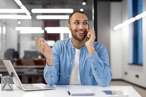 uma homem é falando em dele célula telefone enquanto sentado às uma escrivaninha. ele é sorridente e ele é dentro uma Boa humor foto