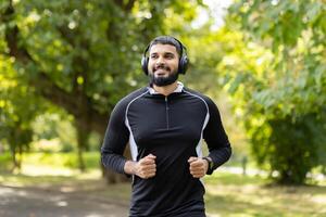 uma em forma jovem homem dentro roupa de esporte é corrida dentro uma ensolarado parque, ouvindo para música com dele fones de ouvido. foto