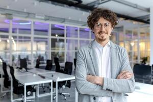 retrato do uma alegre jovem homem de negocios com encaracolado cabelo com confiança em pé dentro uma contemporâneo escritório espaço, braços cruzado. foto