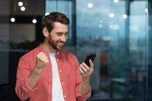 uma bem sucedido homem de negocios alegremente comemora vitória e Boa conquistas, uma homem dentro uma vermelho camisa dentro a escritório recebido a conectados notificação, segurando uma Smartphone dentro dele mãos foto