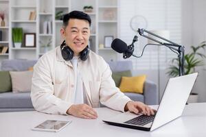 retrato do blogueiro às lar, ásia homem sorridente e olhando às Câmera, homem sentado às mesa com computador portátil dentro vivo sala, gravação audio podcasts e fazendo conectados rádio. foto