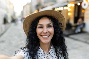 jovem lindo hispânico mulher sorridente e olhando para dentro telefone Câmera, viajante usa Smartphone aplicativo para controlo remoto comunicação chamar, anda em dentro tarde cidade, em turista Tour viagem. foto