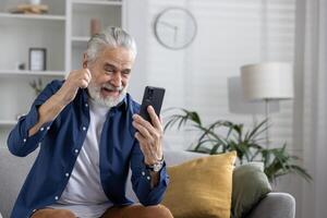 alegre idosos homem alegremente usando uma Móvel telefone, sentado em uma sofá dentro uma bem iluminado vivo quarto com casa decoração. foto