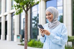 jovem lindo o negócio mulher dentro hijab anda em dentro a cidade lado de fora a escritório prédio, muçulmano mulher usa a inscrição em a telefone, navega conectados Internet Páginas, sorrisos contente. foto
