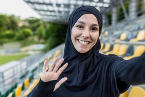 alegre muçulmano mulher dentro hijab levando uma selfie às uma Esportes estádio, acenando e sorridente às a Câmera. foto