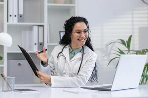 alegre hispânico fêmea médico acionado dentro uma ligar com uma paciente enquanto segurando médico relatórios dentro uma brilhante clínica escritório. foto