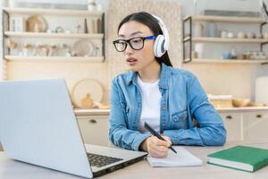 jovem ásia aluna estudando às casa remotamente, mulher assistindo conectados curso sentado dentro cozinha usando computador portátil e bloco de anotações foto