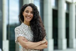 jovem lindo hispânico mulher com encaracolado cabelo lado de fora escritório prédio, companhia trabalhador sorridente e olhando às Câmera com cruzado braços, satisfeito empresária. foto