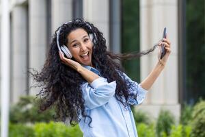 jovem alegre caucasiano mulher com encaracolado cabelo vestindo sem fio fones de ouvido e segurando moderno Smartphone dentro mão ao ar livre cercado de vegetação. fundo do residencial prédio. foto