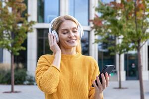 uma alegre meia idade mulher dentro uma amarelo suéter escuta para música em fones de ouvido enquanto segurando uma Smartphone, lado de fora de uma moderno prédio. foto